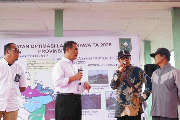 Empat pria berdiri di depan banner kegiatan Optimalisasi Lahan Rawa di Riau. Menteri Pertanian Andi Amran Sulaiman memegang mikrofon dan berdialog dengan petani.